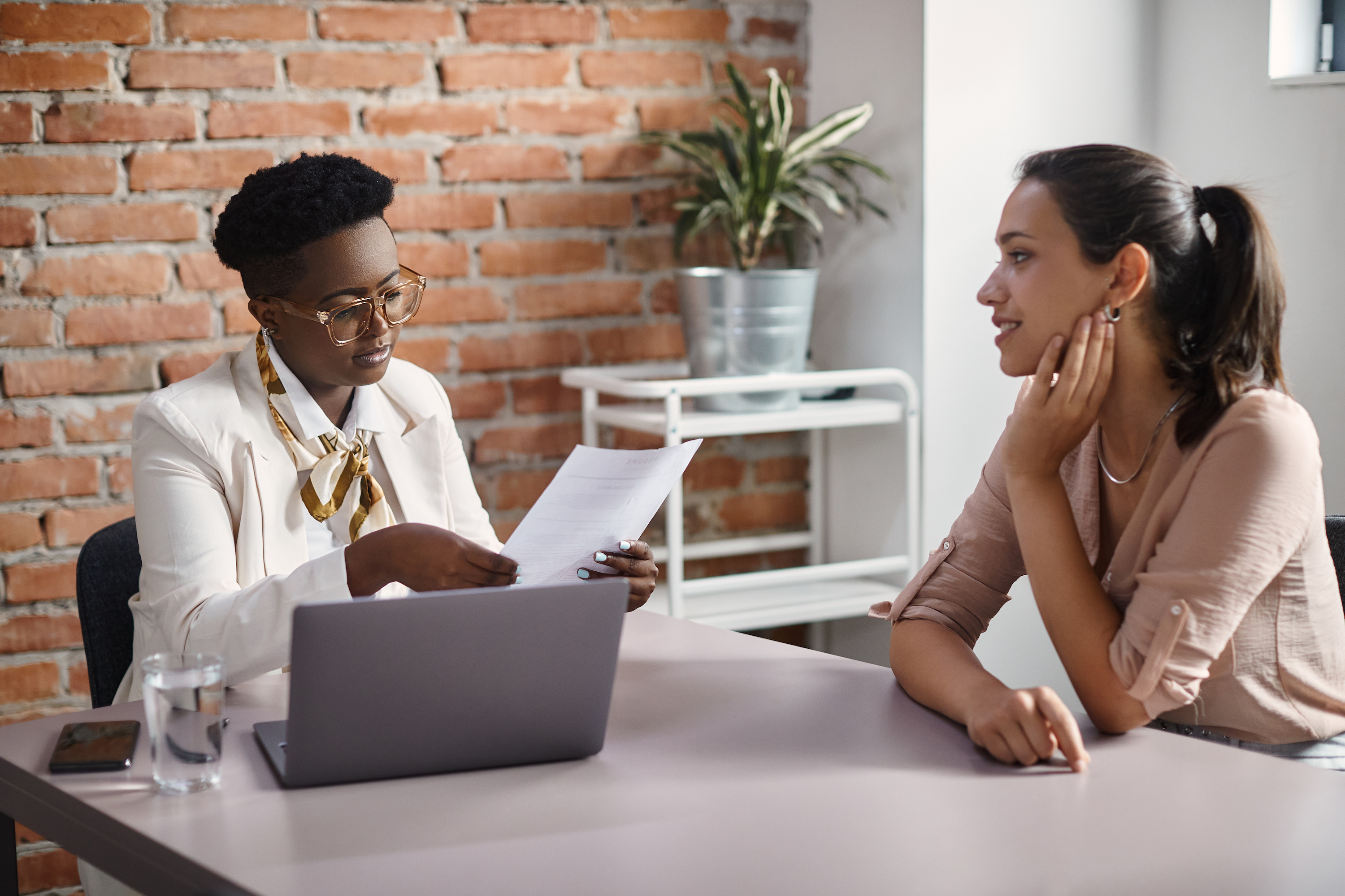 Two people sitting at a table with a laptop talking and reviewing a sheet of paper